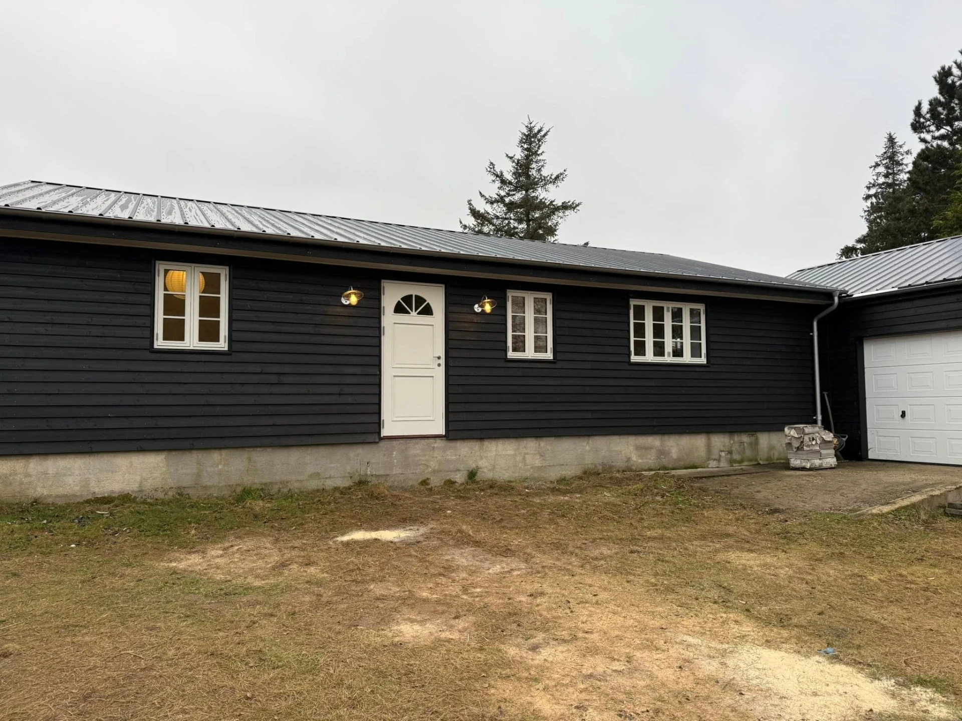 Sort træhus med hvid dør og vinduer, garage, udendørs belysning.